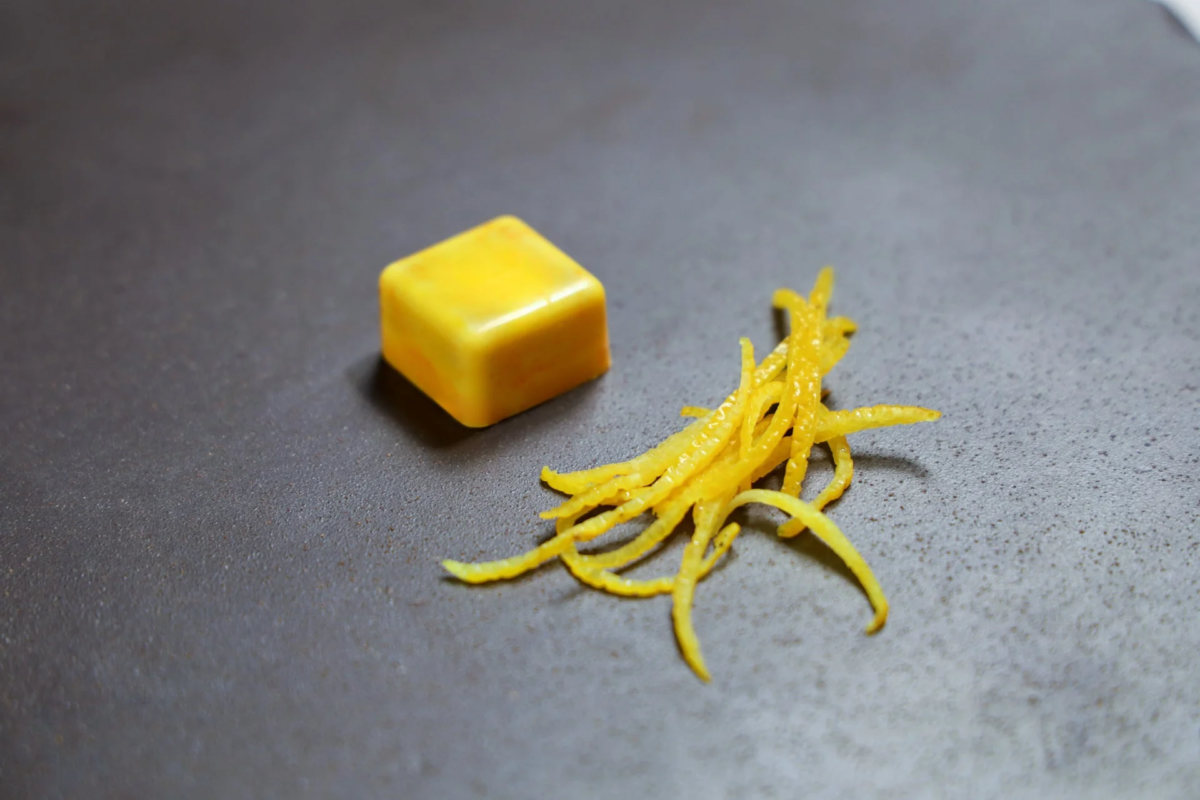 濃いグレーの皿の上に、黄色いキューブ状のデザートと、細く切られた柑橘類の皮（おそらくユズやレモン）が添えられています。シンプルながらも洗練された盛り付けが印象的です。