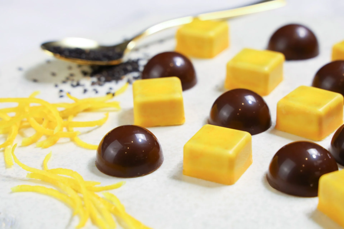 白地の背景に、ドーム型のチョコレートと黄色い四角いチョコレートが並べられています。奥にはポピーシードが乗ったスプーンとレモンピールが添えられており、洗練されたデザートの様子が伺えます。
