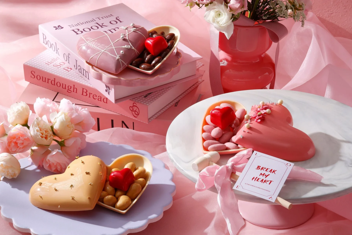 ピンクを基調とした可愛らしい空間に、ハート型のチョコレートやケーキ、花束、そして数冊の本が並べられています。特に目立つのは、中に小さなチョコレートが入ったハート型のスイーツで、ロマンチックな雰囲気が漂っています。バレンタインや特別な日のプレゼントにぴったりの、甘くて魅力的なデザートの盛り合わせです。