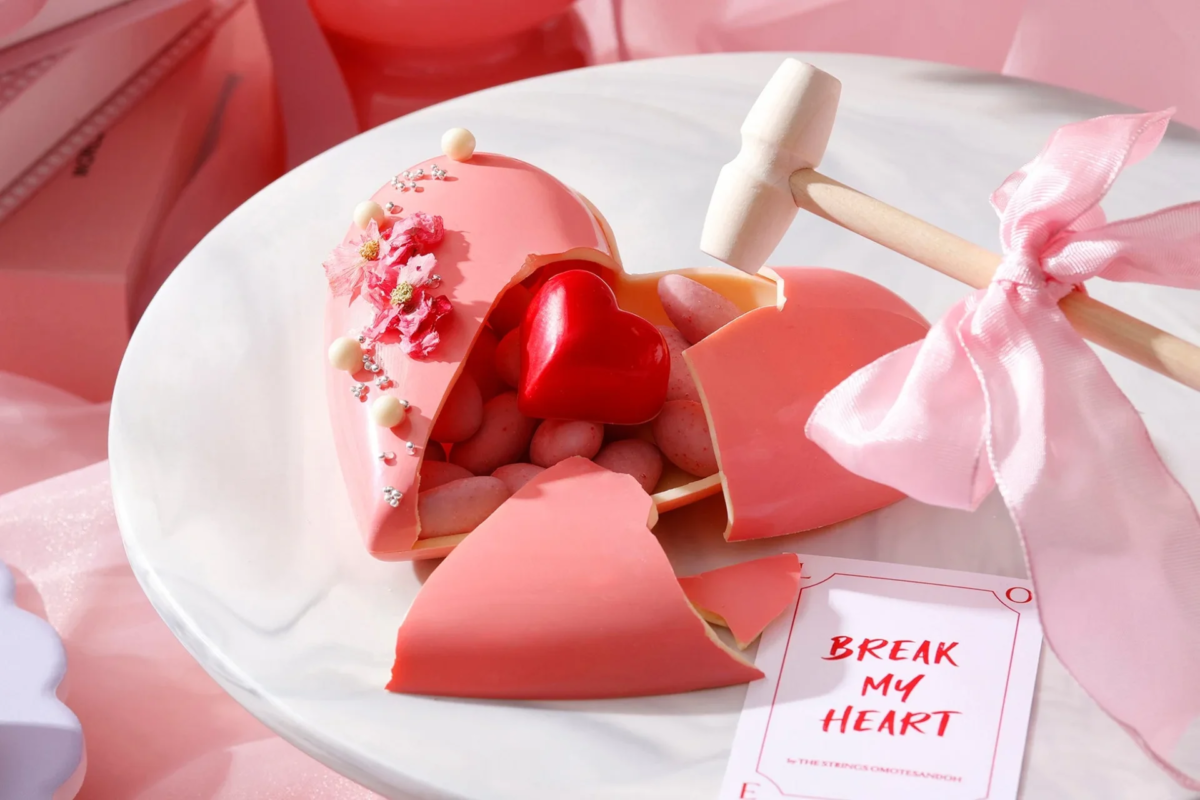 白い皿の上に、ピンクのハート形チョコレートのスマッシュケーキが割れて置かれている。中には小さなキャンディと赤いハートが見え、隣にはピンクのリボンが付いた木製ハンマーがある。バレンタインや記念日にぴったりの、甘くて遊び心のあるギフトを思わせる。