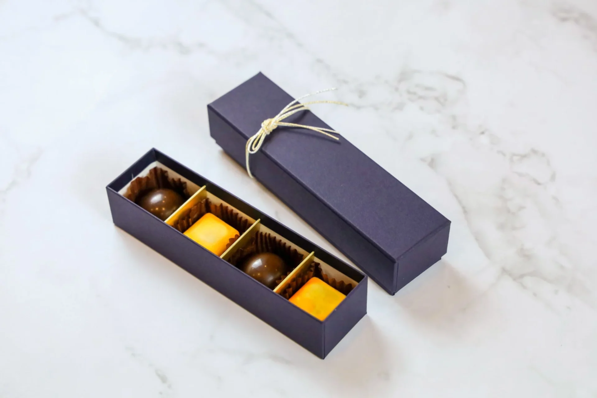 白い大理石のテーブルに置かれた、紫色のギフトボックスに入ったチョコレート。開いた箱には丸いチョコと四角いチョコが2個ずつ並び、もう一つの箱は金色の紐で飾られています。高級感あふれる贈り物です。