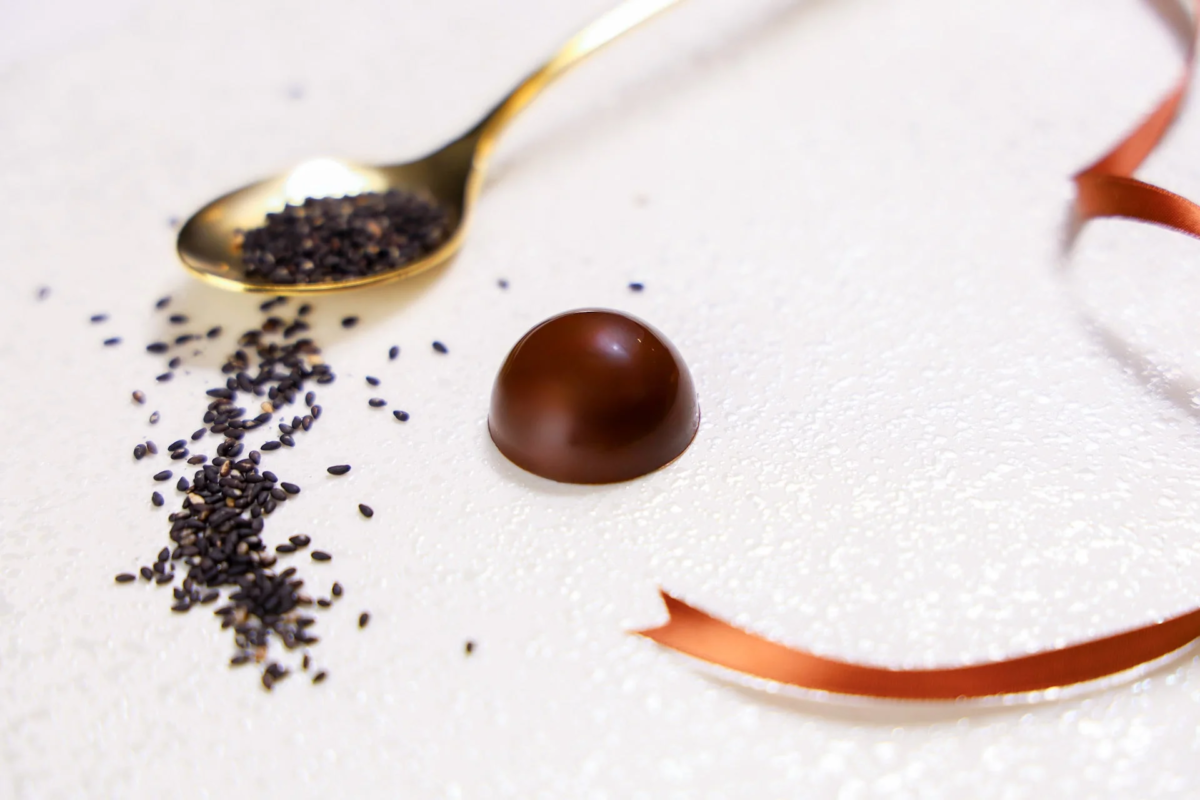 白い背景の上に、光沢のあるチョコレートと、金色のスプーンに乗った黒ごまが散らばっています。茶色のリボンが添えられ、シンプルながらも上品で美味しそうなデザートのイメージです。